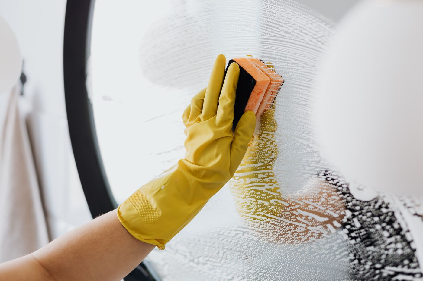 Hände in gelben Handschuhen reinigen einen Badspiegel
